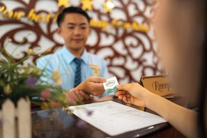 A person handing over a card to a receptionist at a reception desk with a floral arrangement and a decorative wall in the background. - Mali Namphu Hotel (Vientiane)