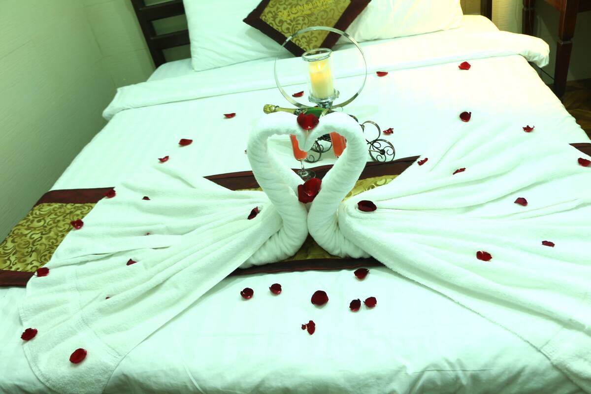 Una cama con una colcha blanca, un arreglo de toallas con forma de corazón y pétalos de rosa esparcidos.