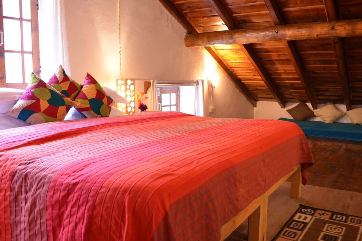 Un dormitorio acogedor con techo de madera, una cama grande con almohadas coloridas y una ventana pequeña.