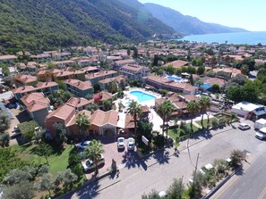Vue aérienne d’une ville côtière, avec un hôtel imposant doté d’une piscine, entouré de bâtiments résidentiels et d’une végétation luxuriante.