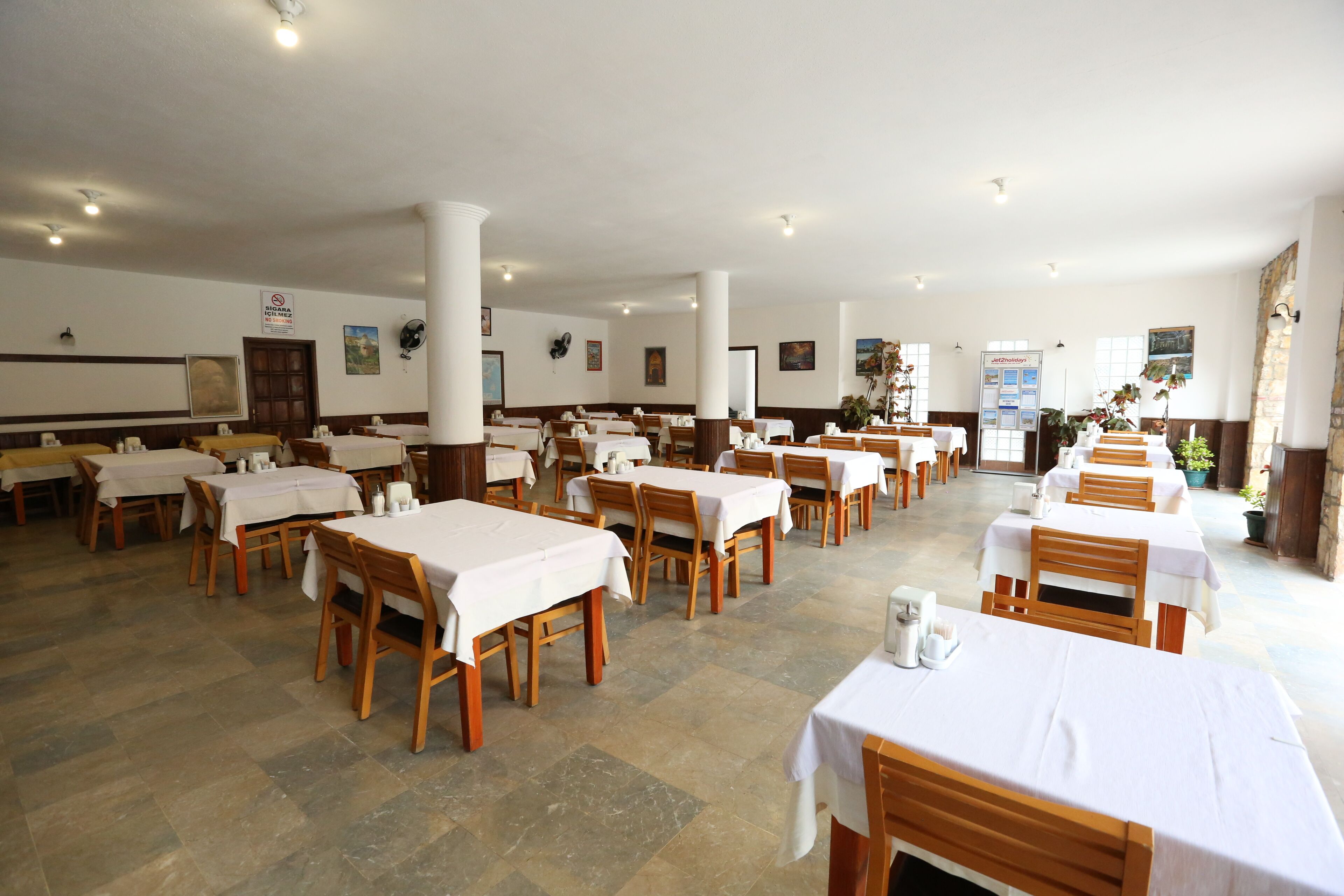 Une salle à manger spacieuse, dotée de nombreuses tables et chaises, de nappes blanches et d’un sol carrelé.