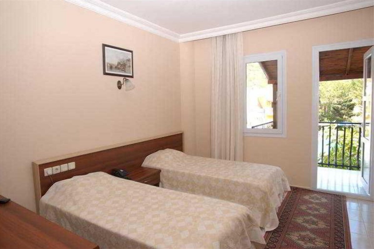 A hotel room with two single beds, a small bedside table, a window with curtains, and a view of the outdoors.