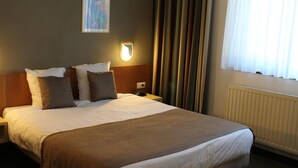 Une chambre d’hôtel avec un grand lit, une table de chevet, une lampe fixée au mur, une fenêtre avec des rideaux et un radiateur.