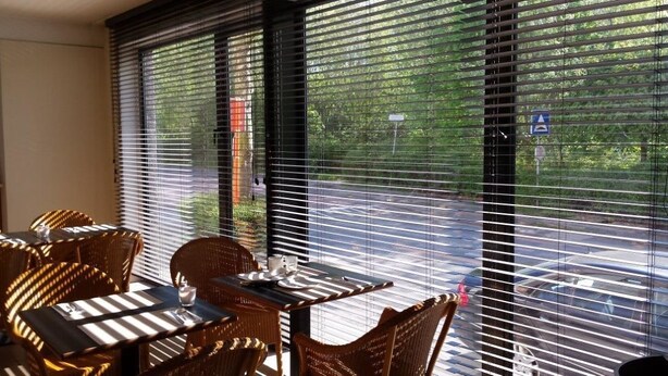 Un coin repas avec des chaises et des tables en osier, de grandes fenêtres équipées de stores et une vue sur la rue.