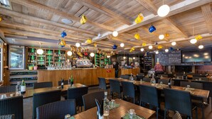 A restaurant with wooden ceiling beams, hanging light fixtures, and a bar area with shelves of bottles and glasses. Tables are set with cutlery and napkins. - Logis Hôtel l'Aubrac (Laguiole)
