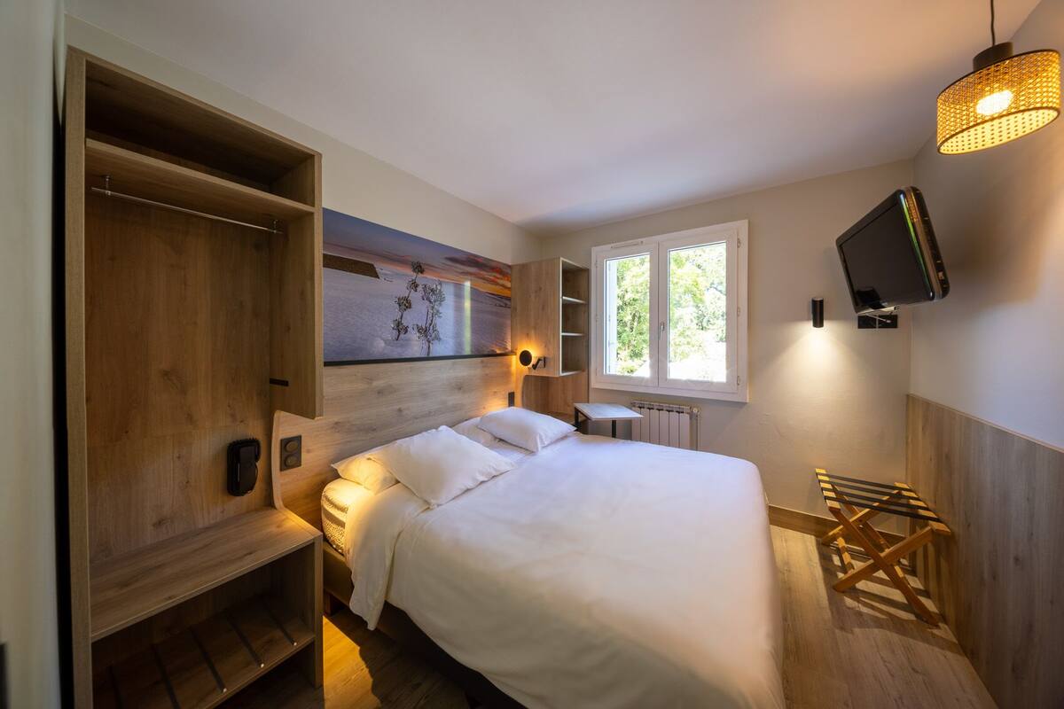 A bedroom with a large bed, wooden shelving, a TV mounted on the wall, and a window with a view of greenery.