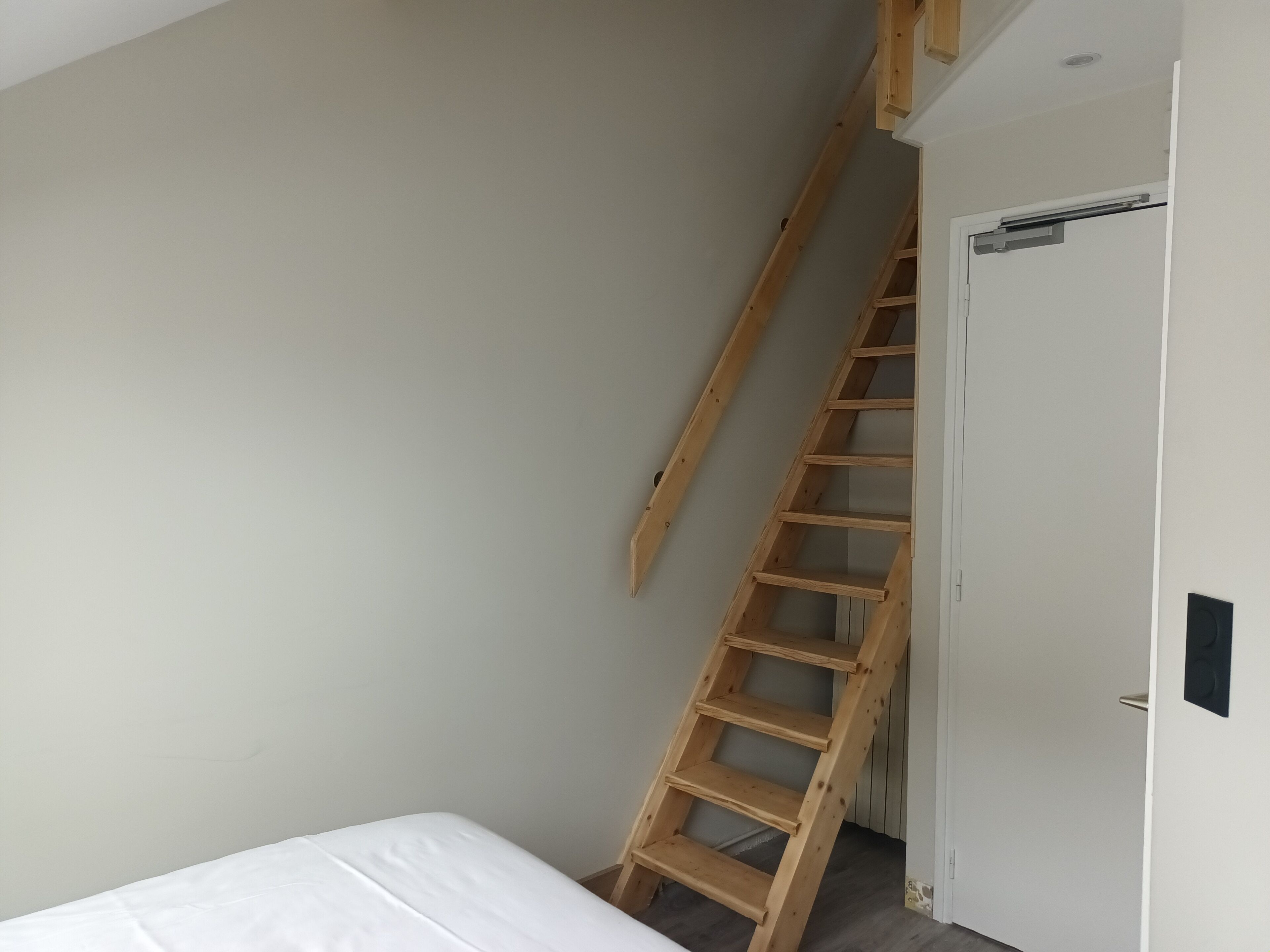 Een slaapkamer met een houten ladder naar een deur, een wit bed en een schuine plafond.