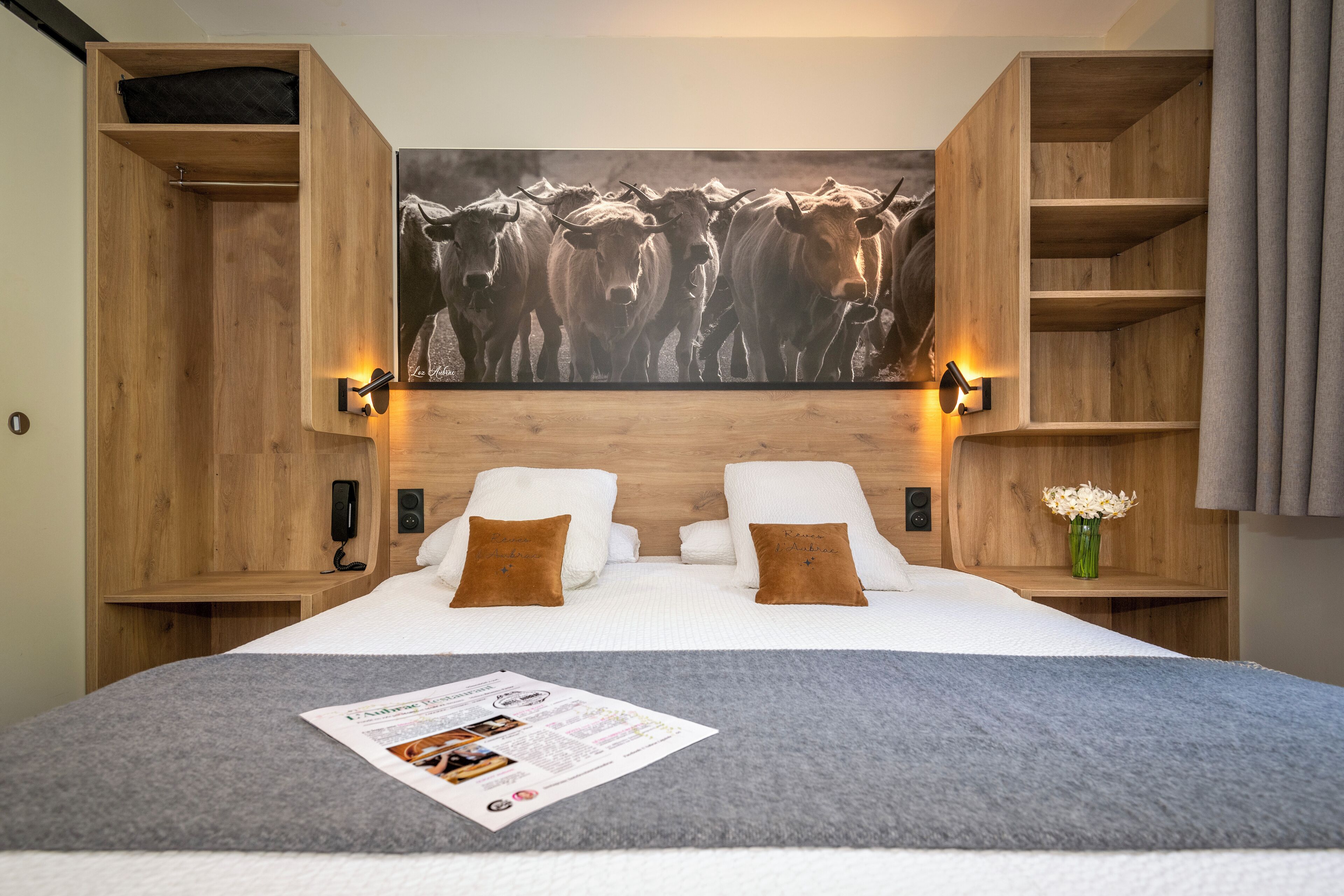 Quarto de hotel com uma cama grande, duas mesas de cabeceira, uma cabeceira de madeira, um quadro fixado na parede com gado e um abajur.