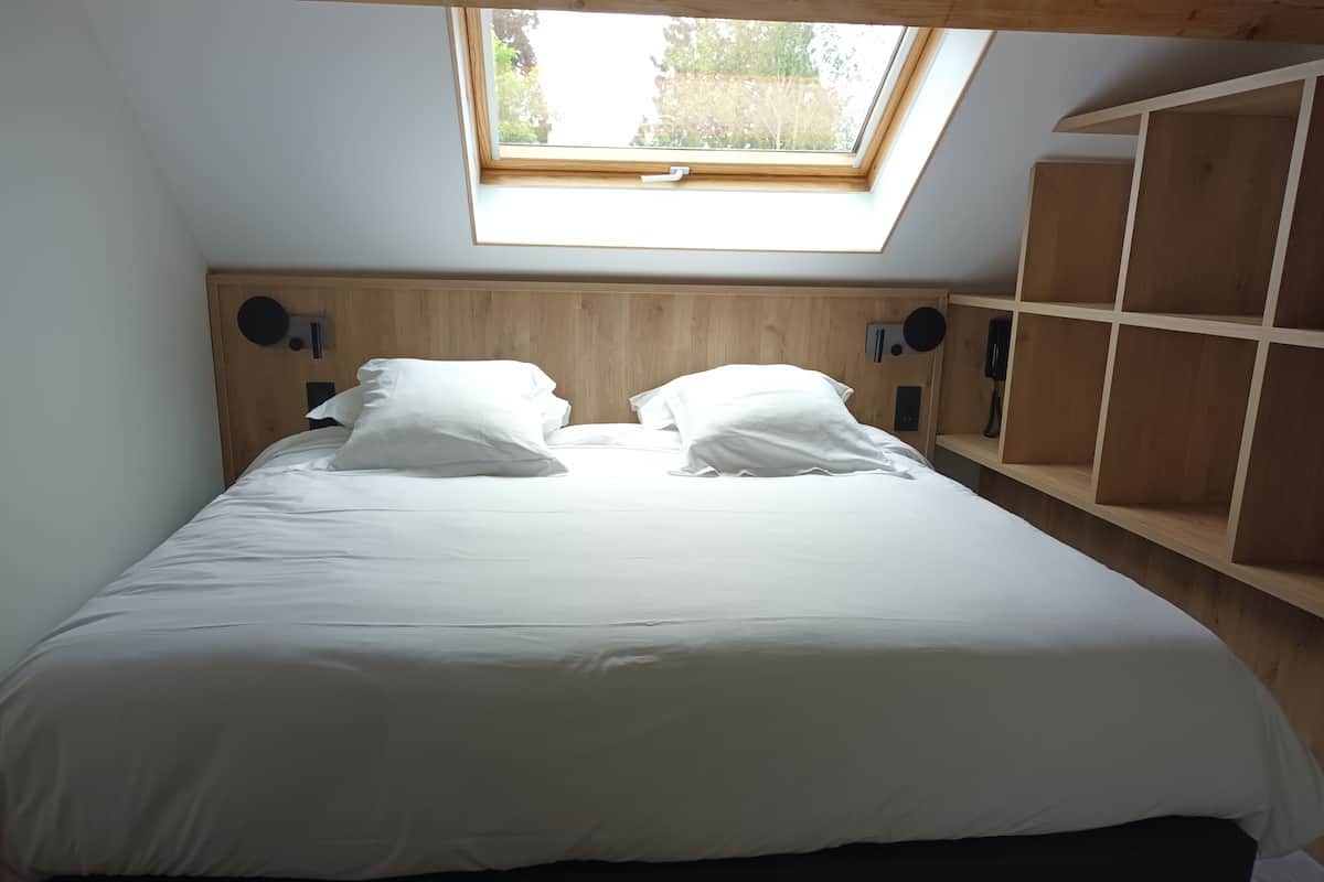 A bedroom with a skylight, a bed with white bedding, and wooden headboard and shelves.