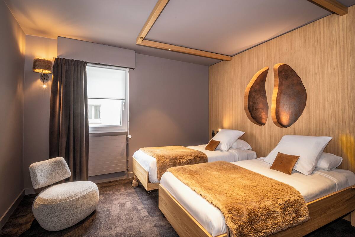 A room with two beds, a chair, a window, and a wall-mounted light fixture.
