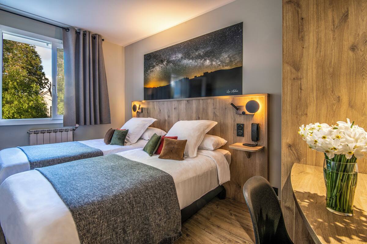 A hotel room with two beds, a night sky mural, a wooden headboard, a bedside table with a lamp, and a window with curtains.