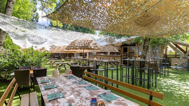 An outdoor dining area with tables and benches, surrounded by greenery and thatched-roof structures. - Logis Hôtel l'Aubrac (Laguiole)