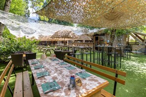An outdoor dining area with tables and benches, surrounded by greenery and thatched-roof structures. - Logis Hôtel l'Aubrac (Laguiole)