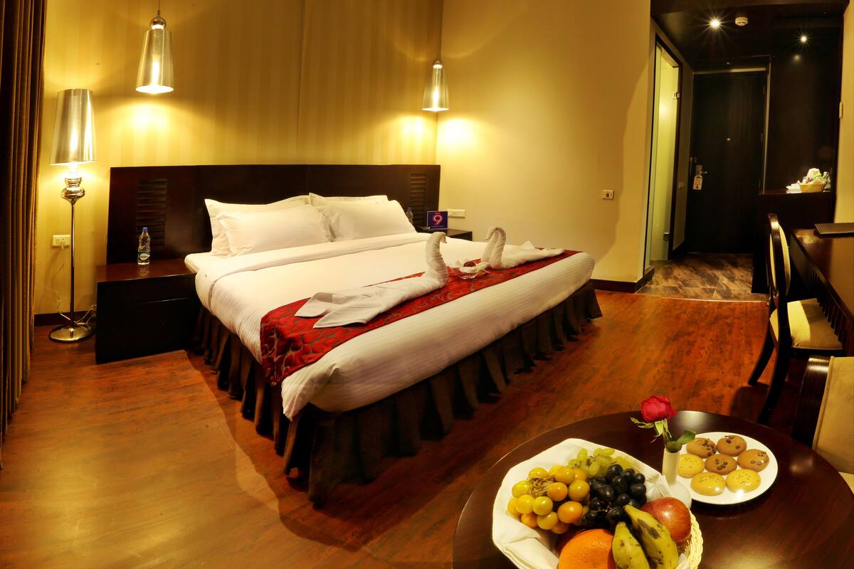 A hotel room with a neatly made bed, a dining table with fruit and a rose, and a view of the hallway.