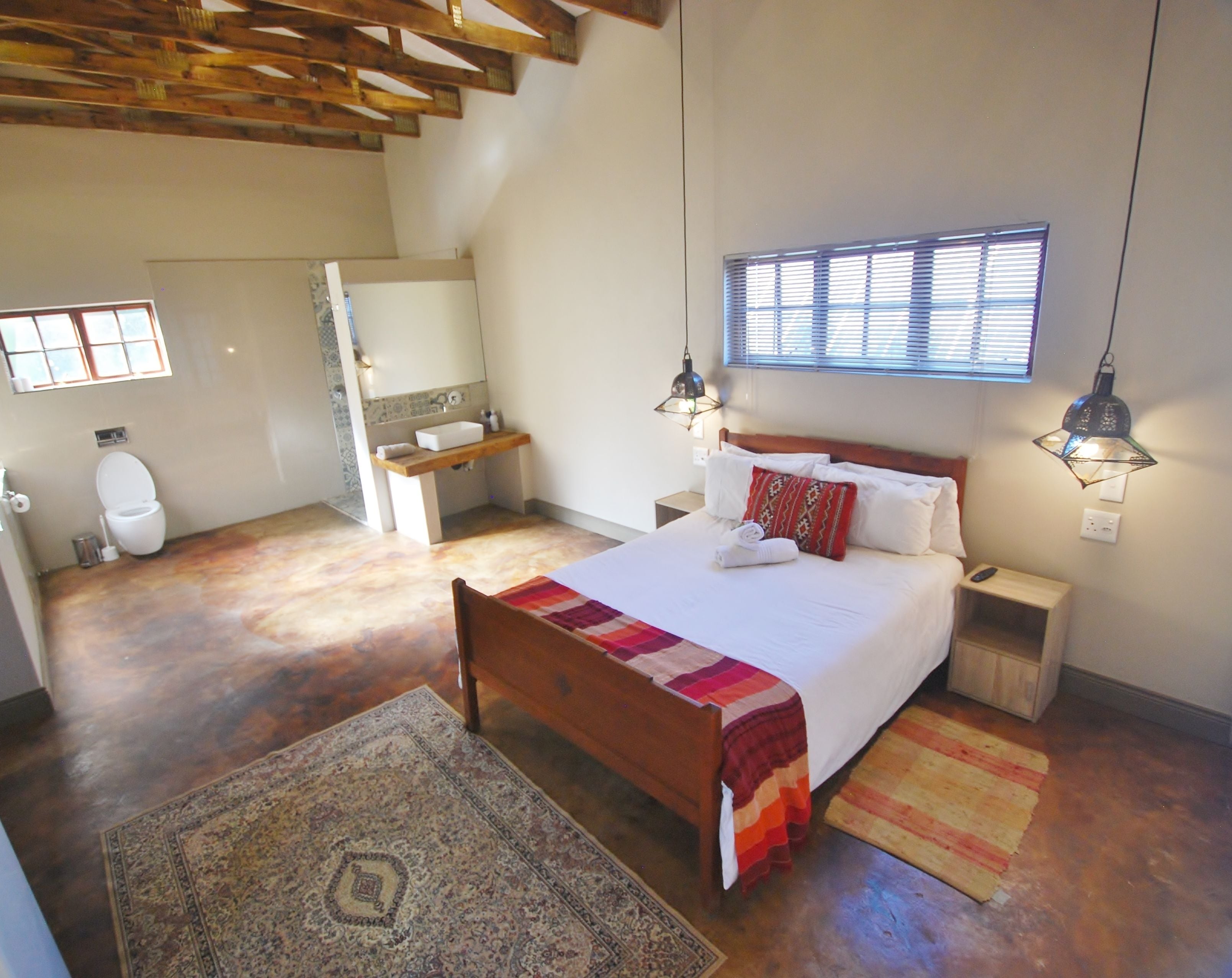Une chambre à coucher avec un lit en bois, une table de chevet, une commode, une fenêtre avec des stores, une salle de bain avec des toilettes, un lavabo et un miroir.