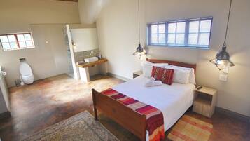Une chambre à coucher avec un lit en bois, une table de chevet, une commode, une fenêtre avec des stores, une salle de bain avec des toilettes, un lavabo et un miroir.