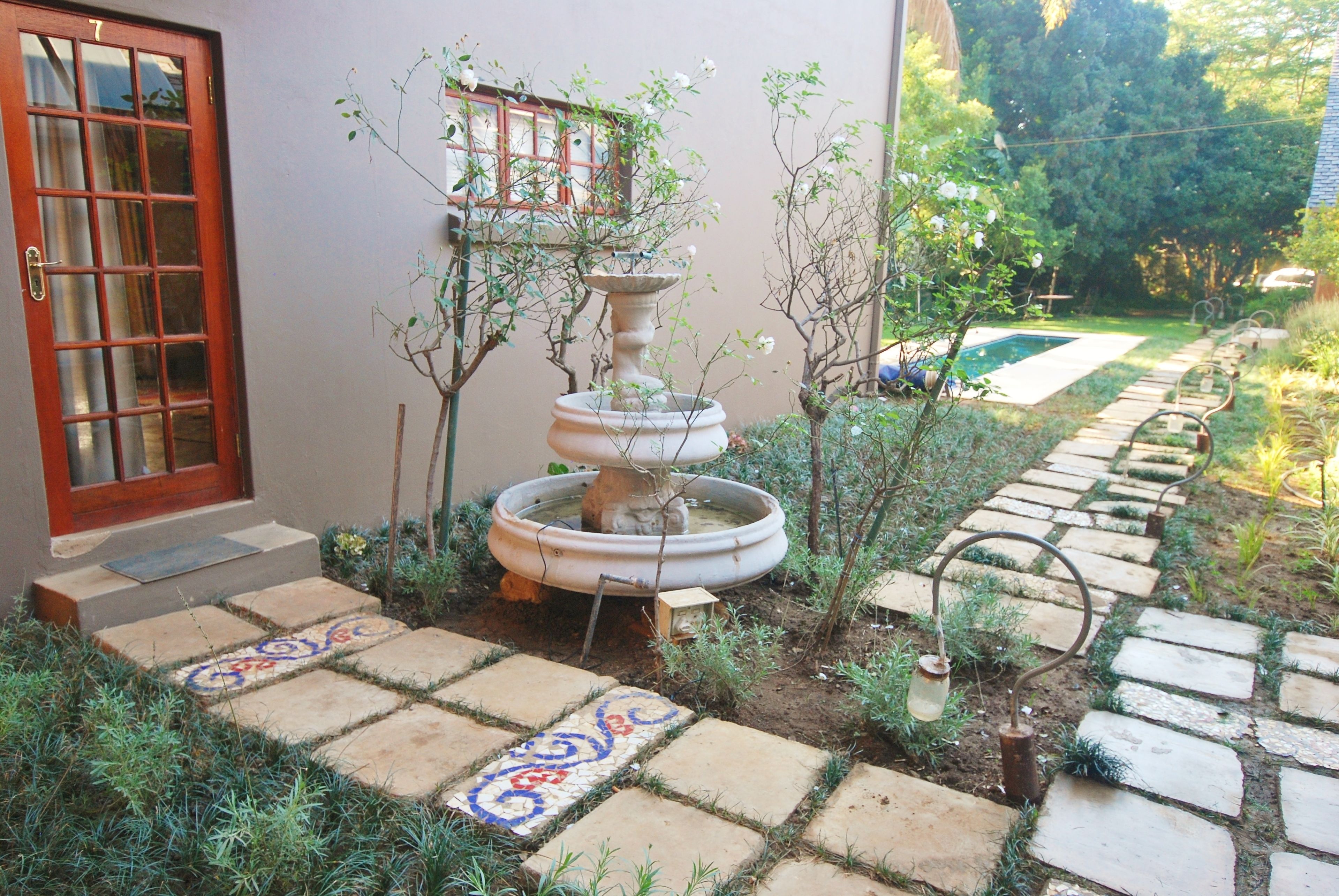 A garden with a stone pathway leading to a door, a small fountain, and a pool in the background.