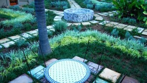 Ein Garten mit einem runden Tisch, umgeben von quadratischen Fliesen, einem Kiesweg und verschiedenen Pflanzen.