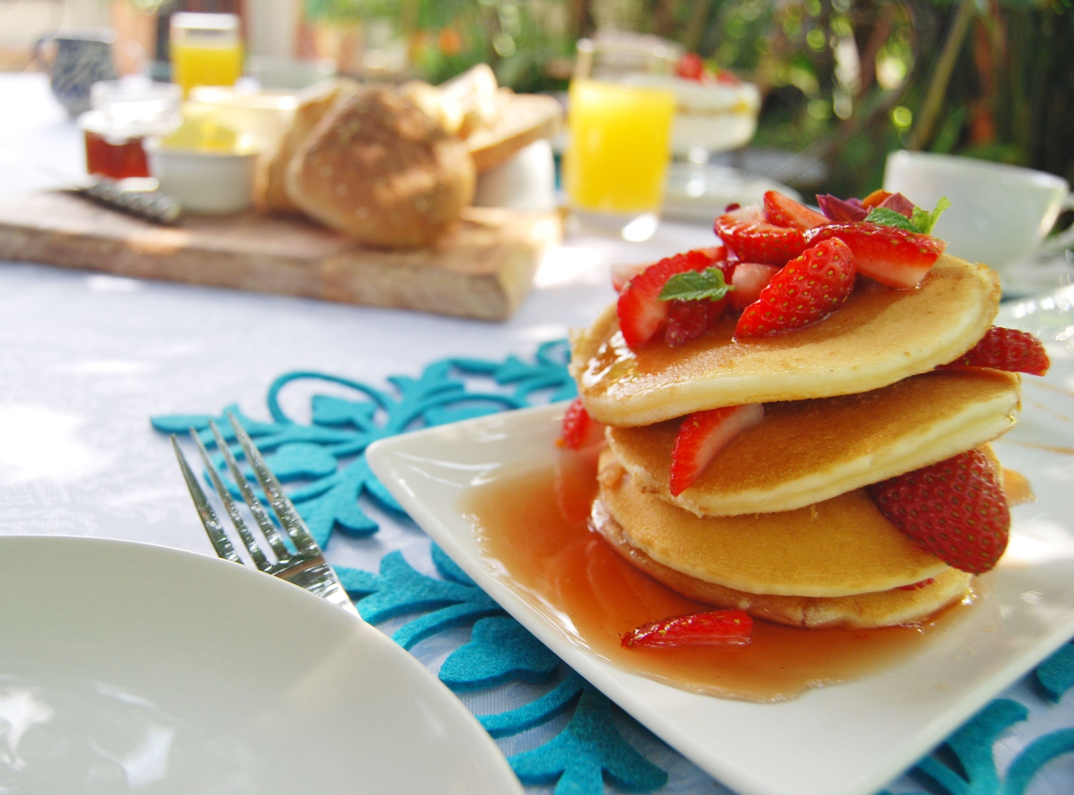 A breakfast setting with pancakes topped with strawberries and syrup, a croissant, and a glass of orange juice on a table with a blue tablecloth.