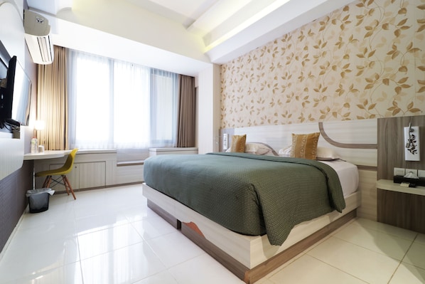 Relax in a serene space with a large bed, bedside table, and desk, surrounded by a leaf pattern wallpaper and natural light.