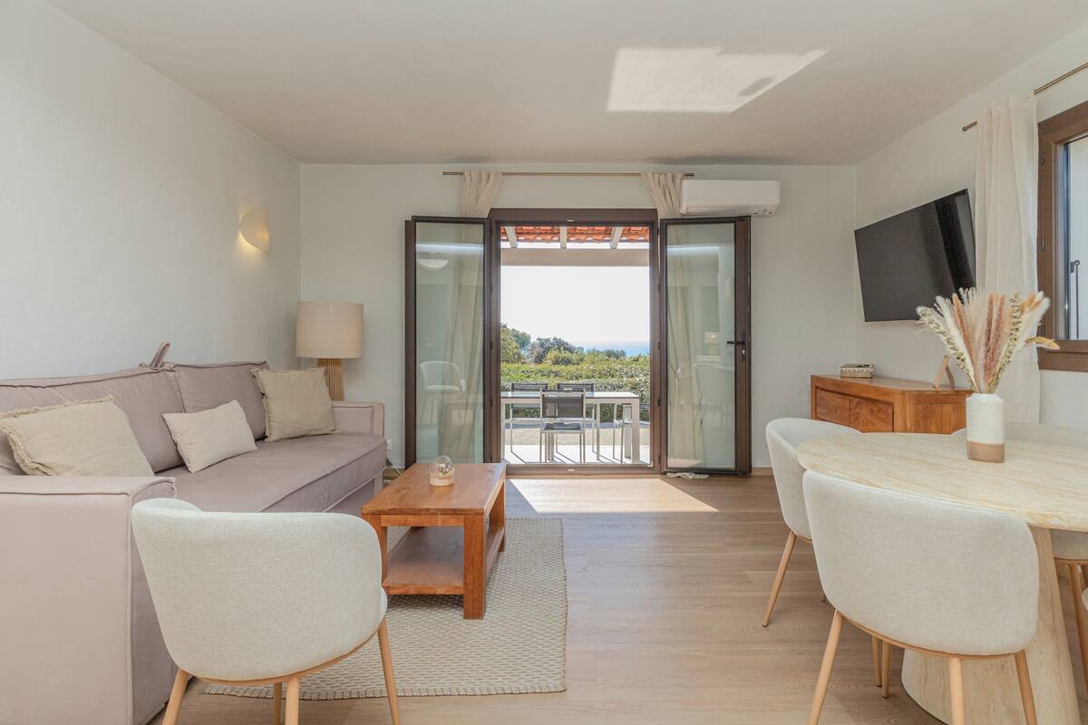 A modern living room with a sofa, coffee table, and chairs, a dining table with chairs, a TV mounted on the wall, and a sliding glass door leading to an outdoor area.