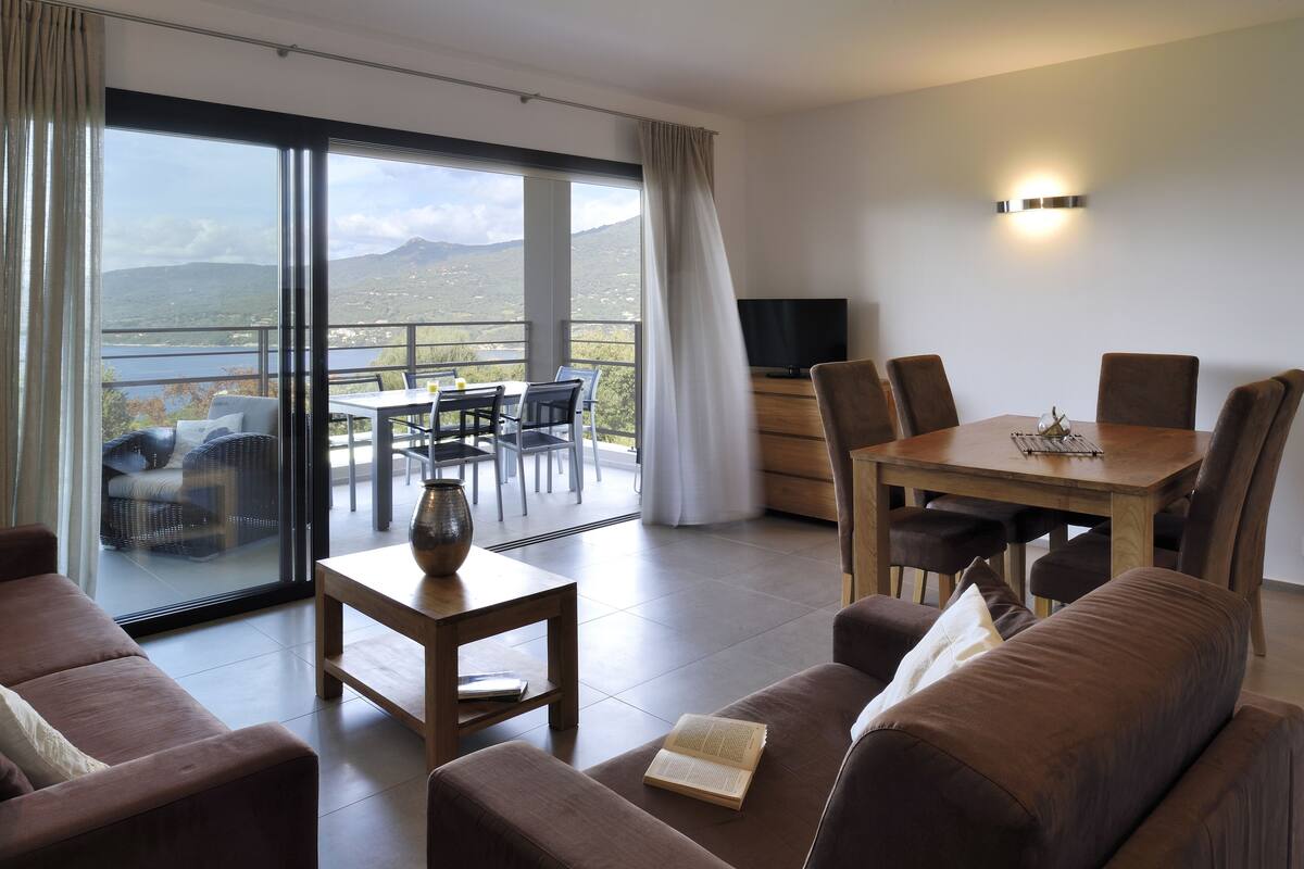 A modern living room with a sofa, a coffee table, and a dining area with a table and chairs. A TV is mounted on the wall, and there is a large window with a view of mountains and a lake.
