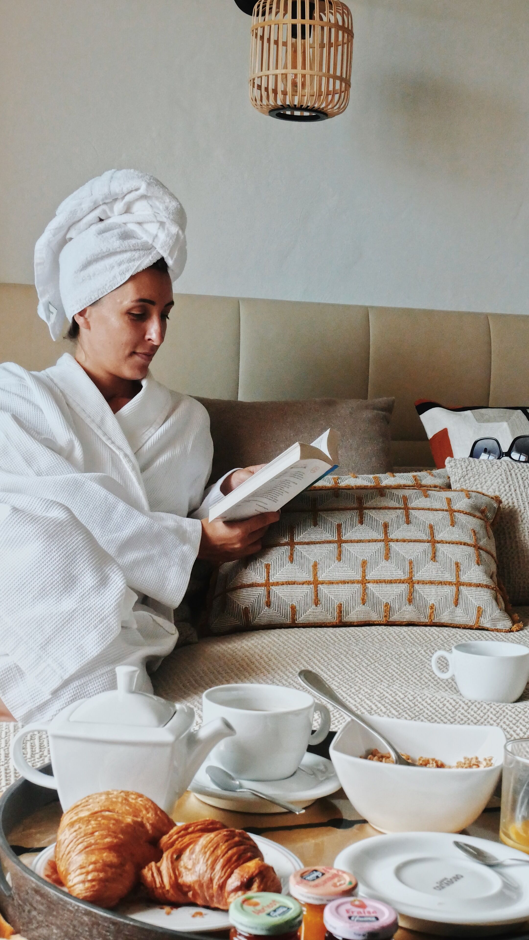 A person reading a book in a room with a bed, pillows, a tray with croissants, cups, and a teapot.