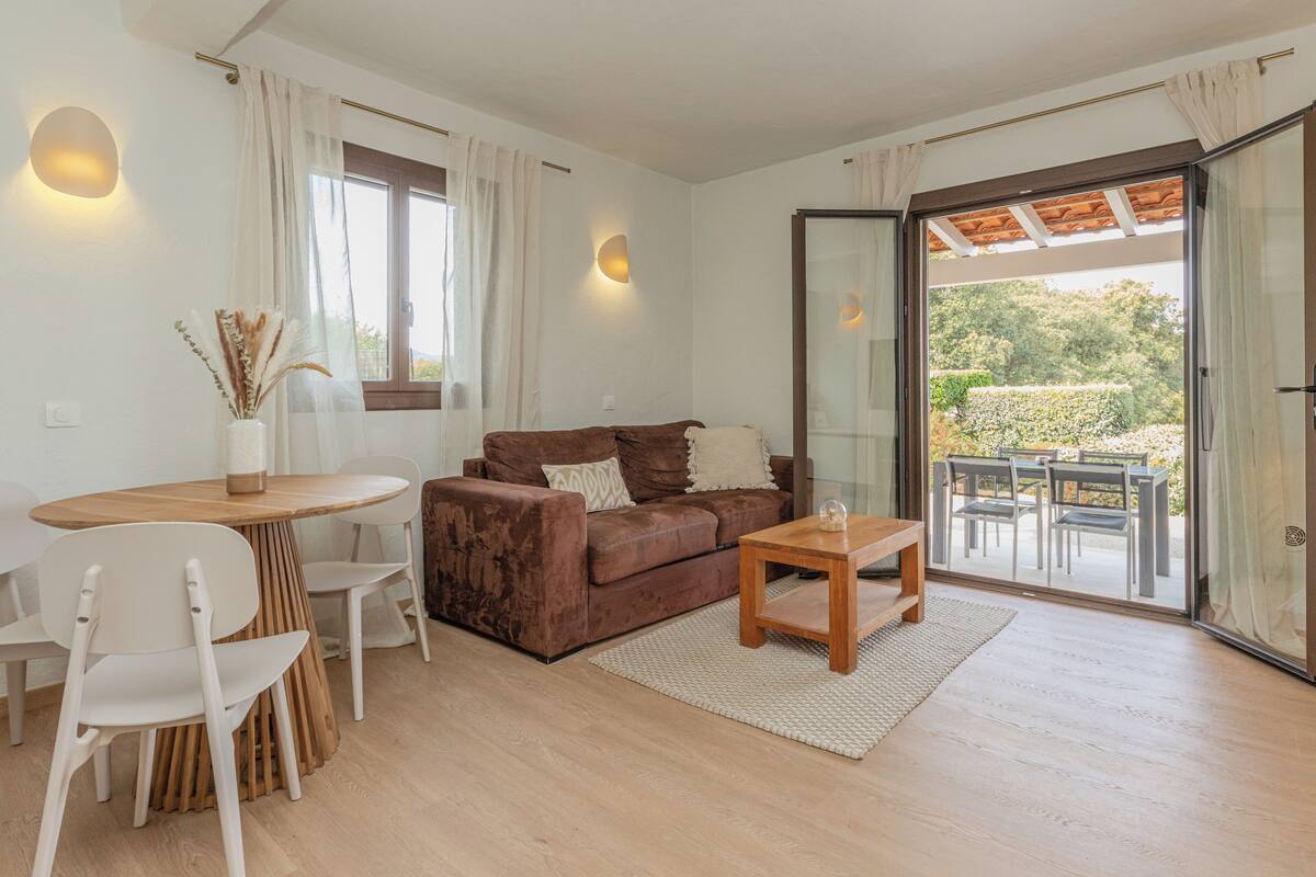 A living room with a brown sofa, white chairs, a wooden coffee table, and a glass door leading to an outdoor area.