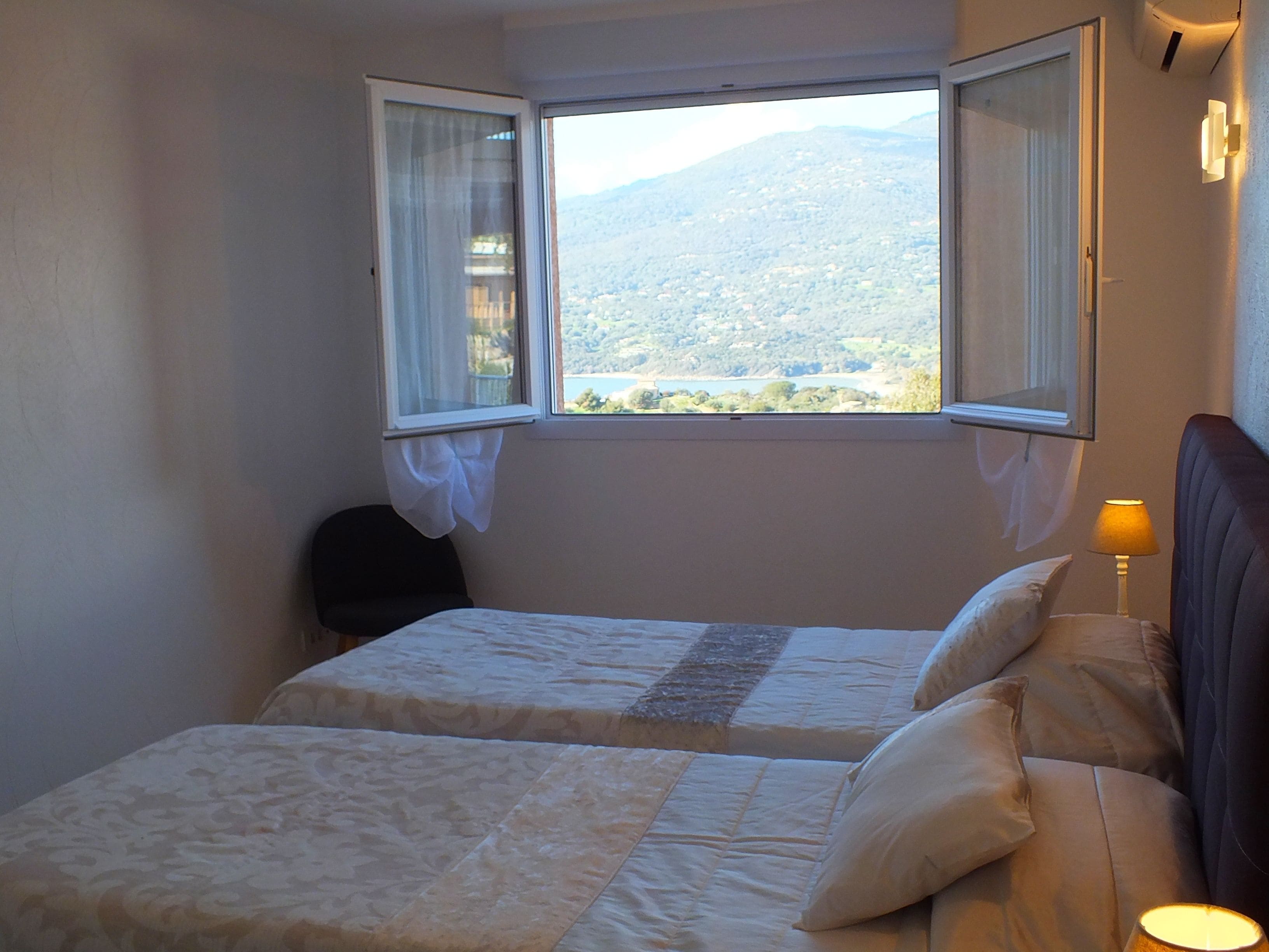 A room with two beds, a window with curtains, a lamp, and a view of a mountainous landscape.