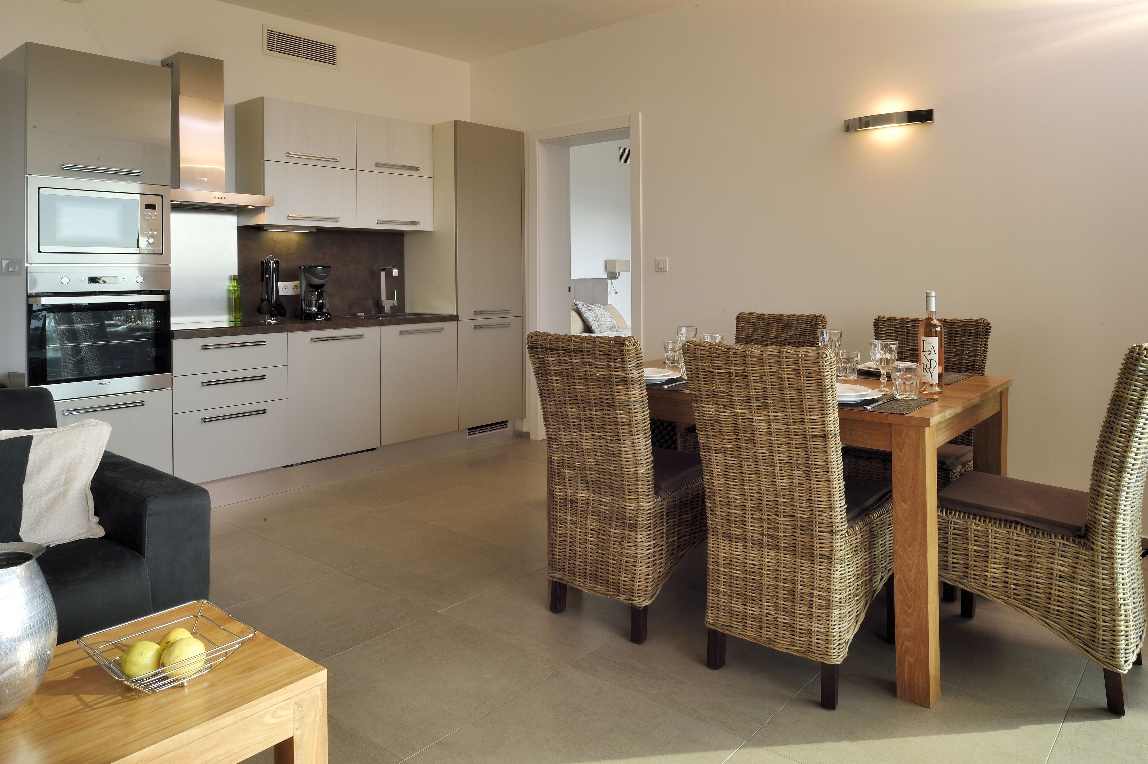 A modern kitchen with built-in appliances, a dining area with wicker chairs, and a small seating area with a table and cushions.