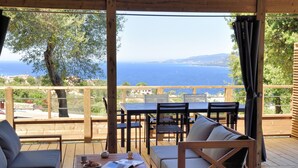 Une terrasse en bois avec un espace salon extérieur, comprenant un canapé et des fauteuils, offrant une vue panoramique sur la mer et les collines.