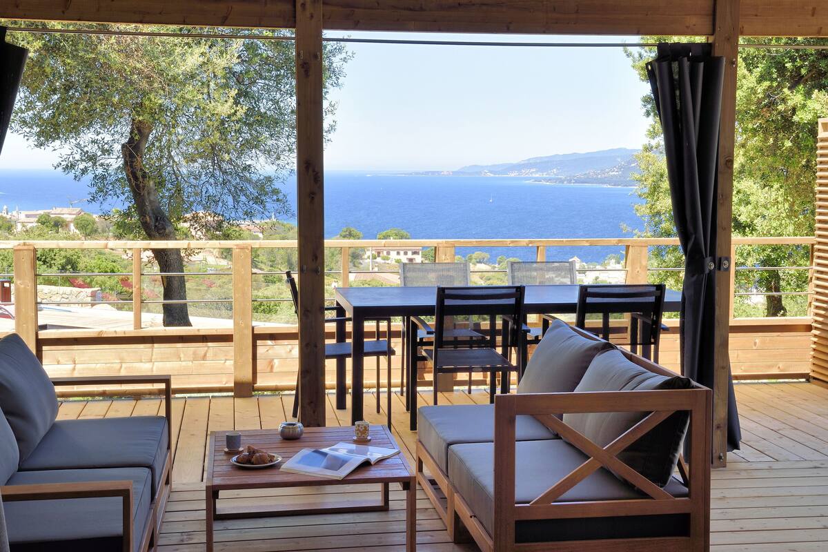 A wooden deck with outdoor seating, including a sofa and chairs, overlooking a scenic view of the sea and hills.