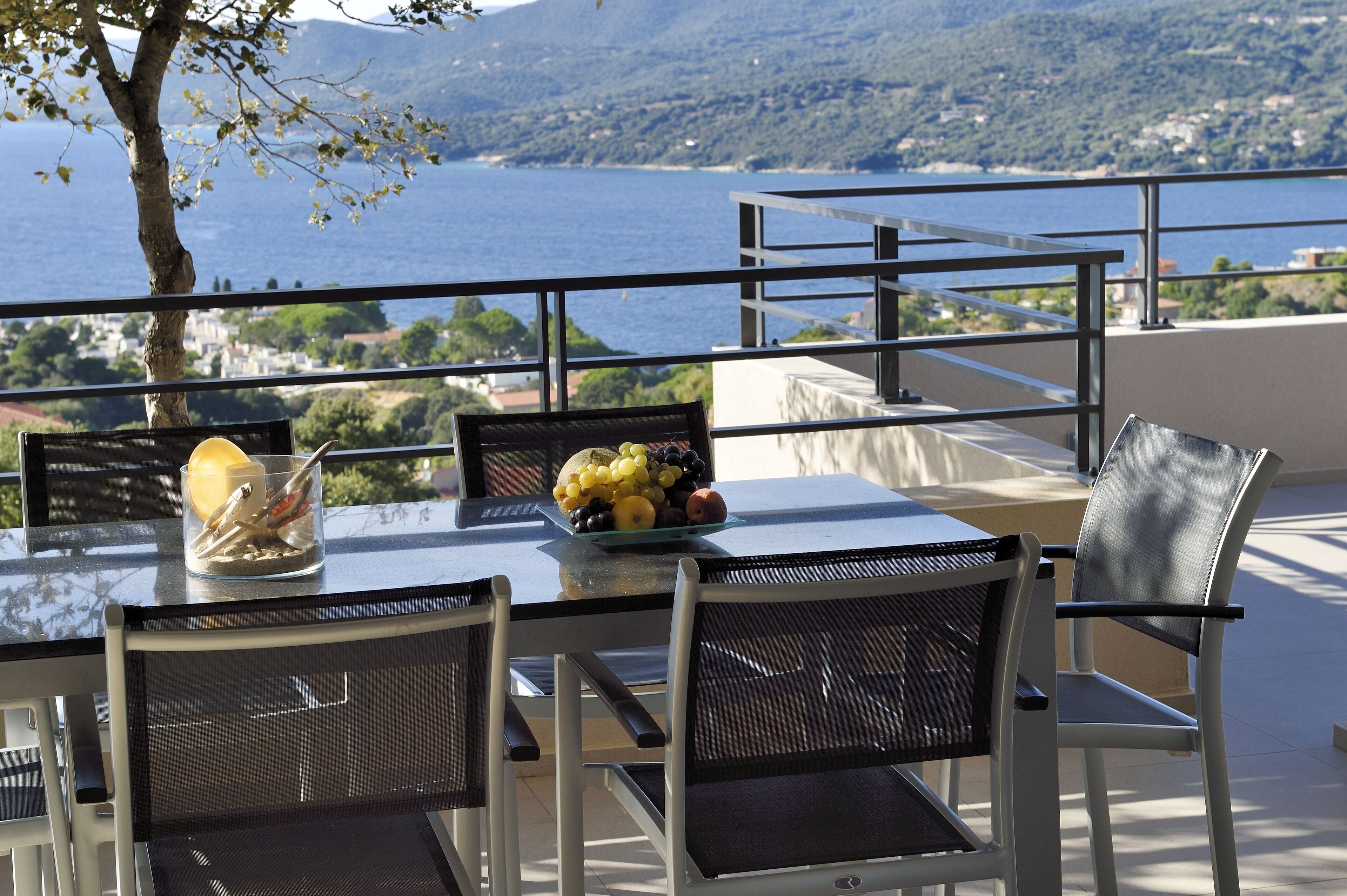 Ein Tisch mit Obstschale und einem Glas Wasser auf einem Balkon mit Blick auf ein Gewässer und eine Berglandschaft.
