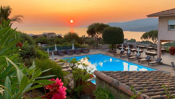 Un espace piscine doté de chaises longues et de tables, entouré de verdure et offrant une vue sur un plan d’eau avec des montagnes en arrière-plan.