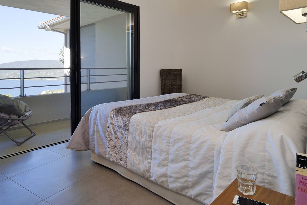 A bedroom with a bed, a bedside table with a glass and a book, a chair, and a view of the sea through a sliding door.