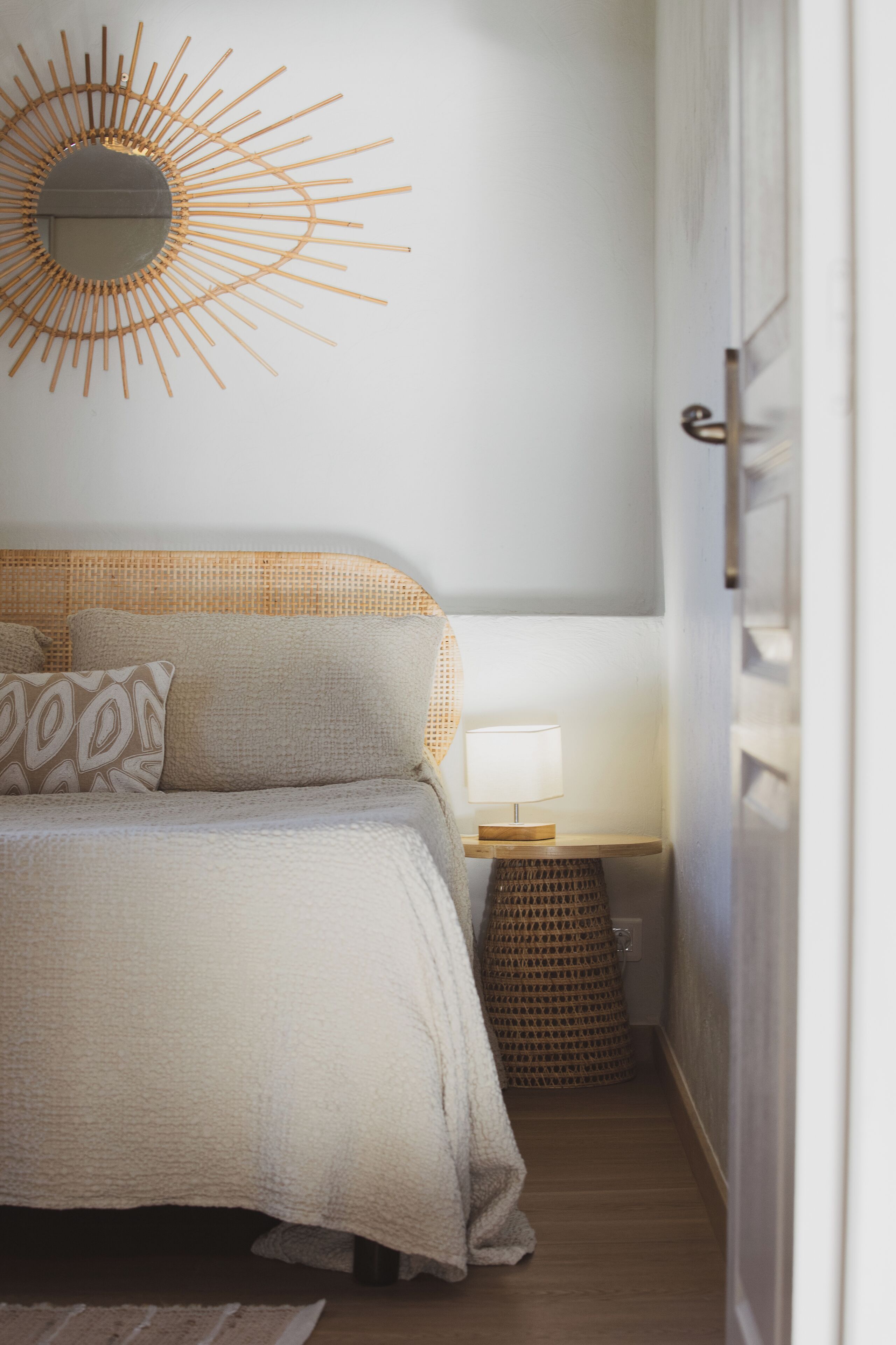 Un dormitorio con una cama, un espejo adosado a la pared, una mesita de noche con lámpara y una cesta tejida.