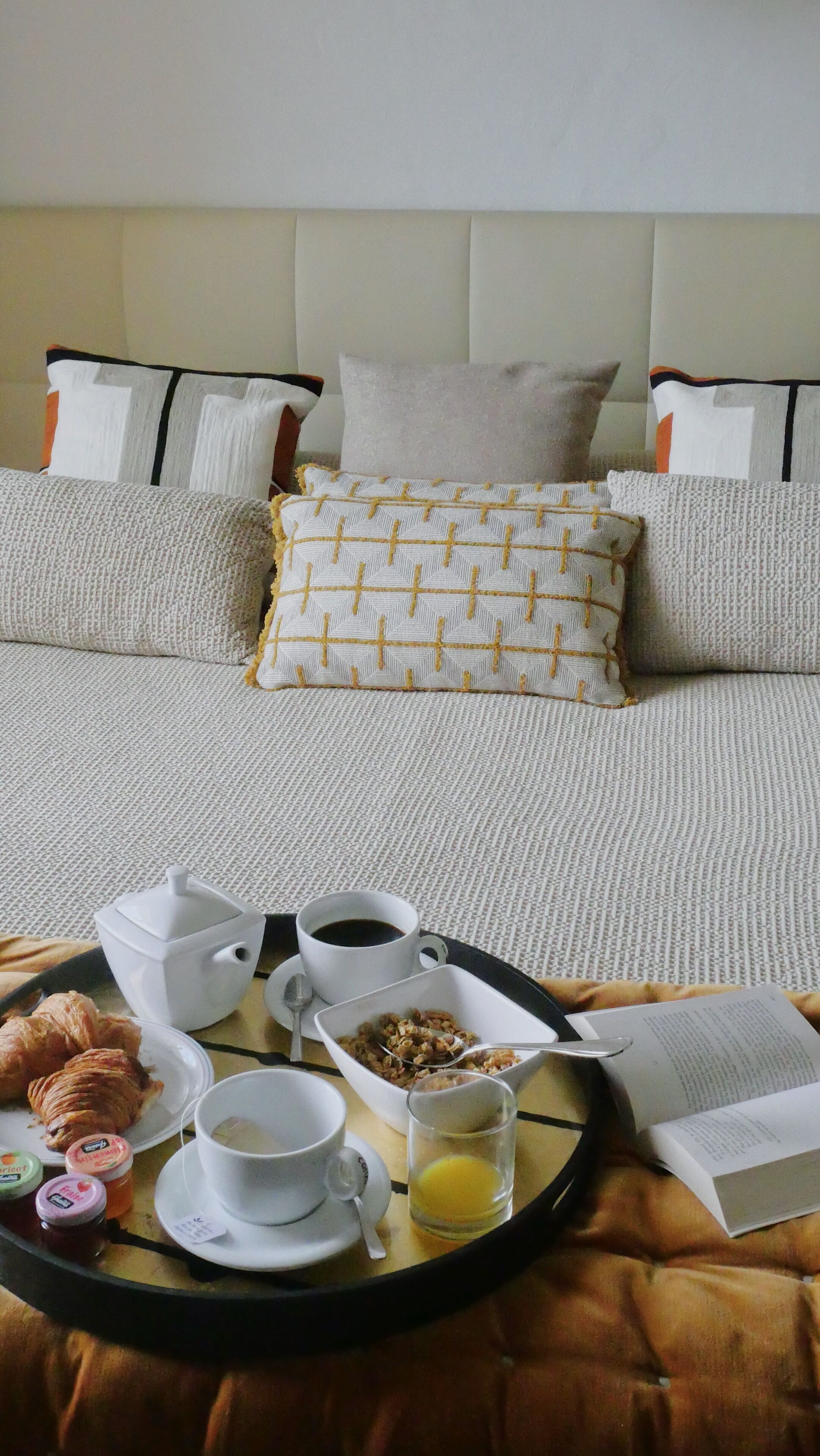 A neatly made breakfast tray with croissants, coffee, cereal, and juice on a bed with a headboard and pillows.