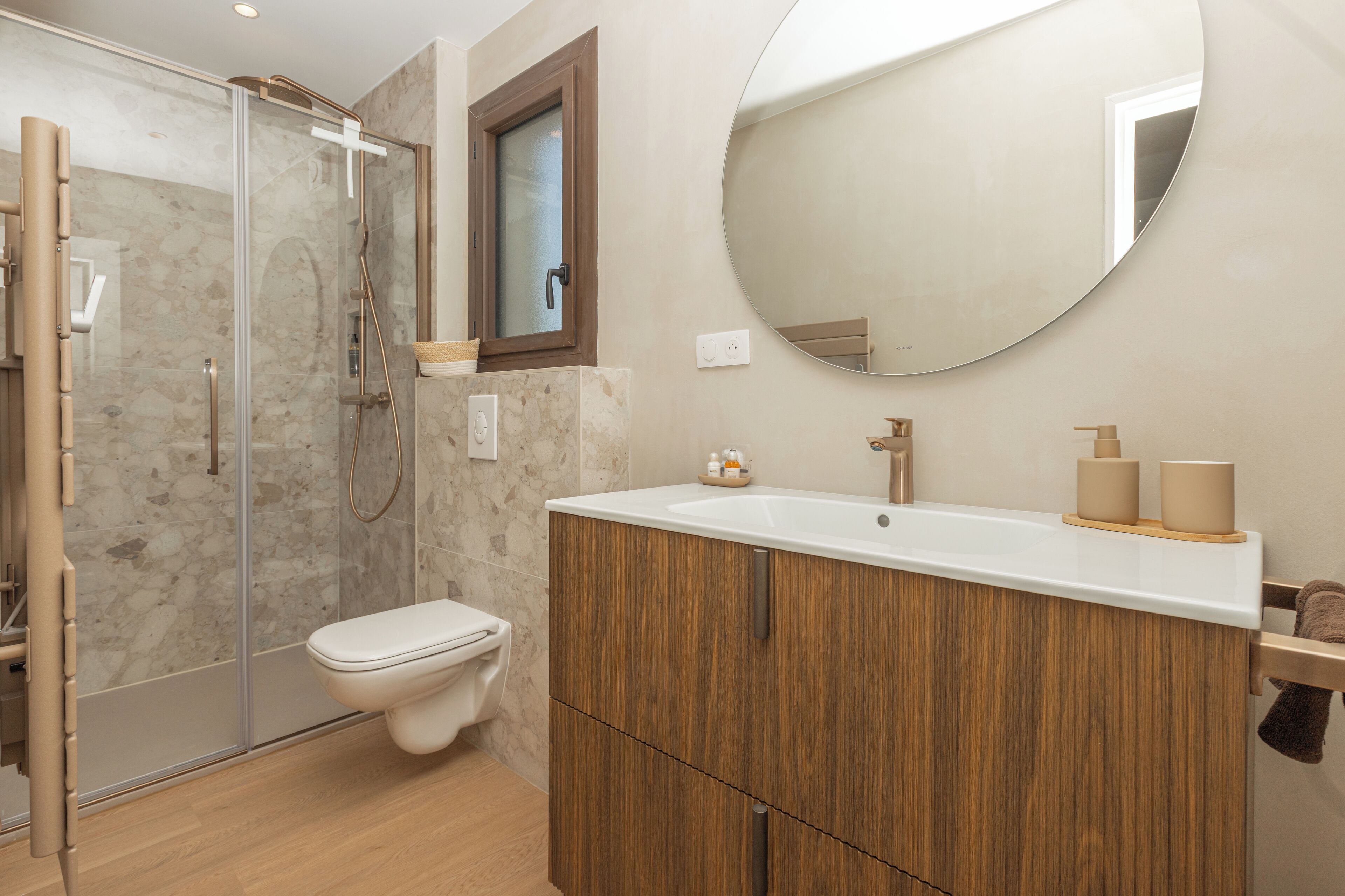 Een moderne badkamer met een glazen douchecabine, een houten wastafelmeubel en een ronde spiegel.