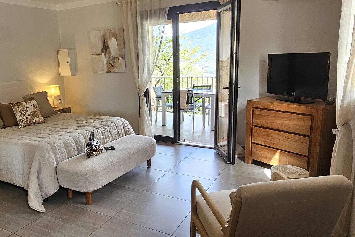 A bedroom with a bed, a TV, a wooden dresser, and a balcony with a view.