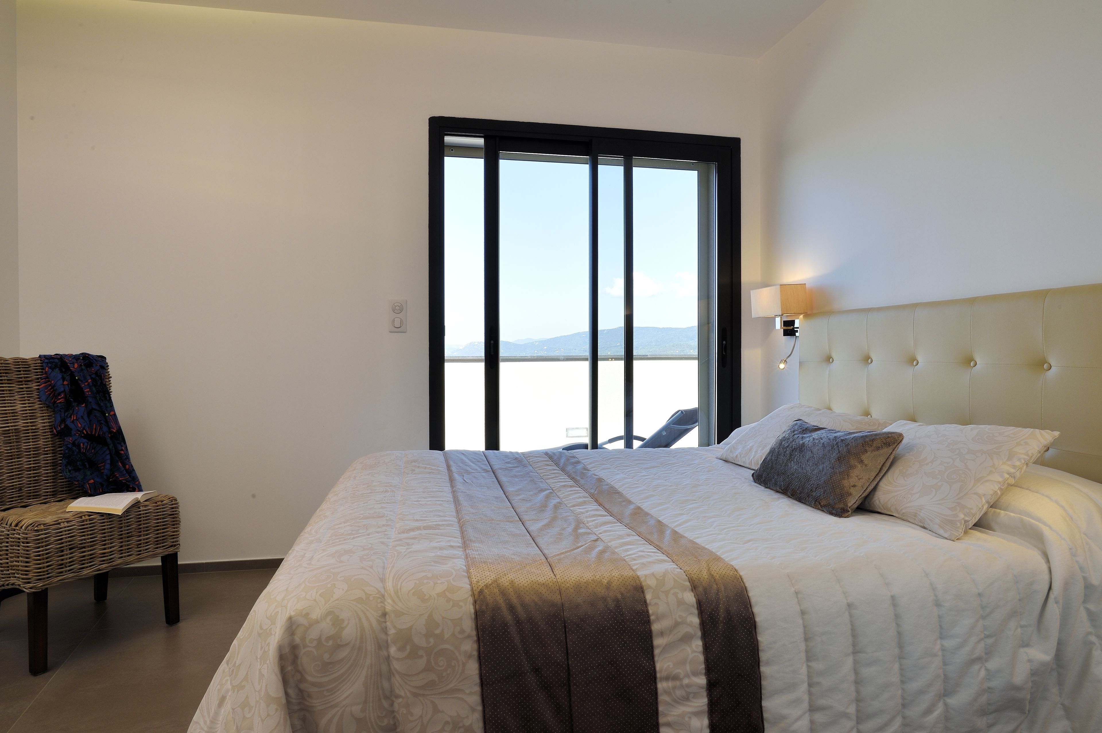 A bedroom with a large bed, a chair, and a view of the sea through a window.