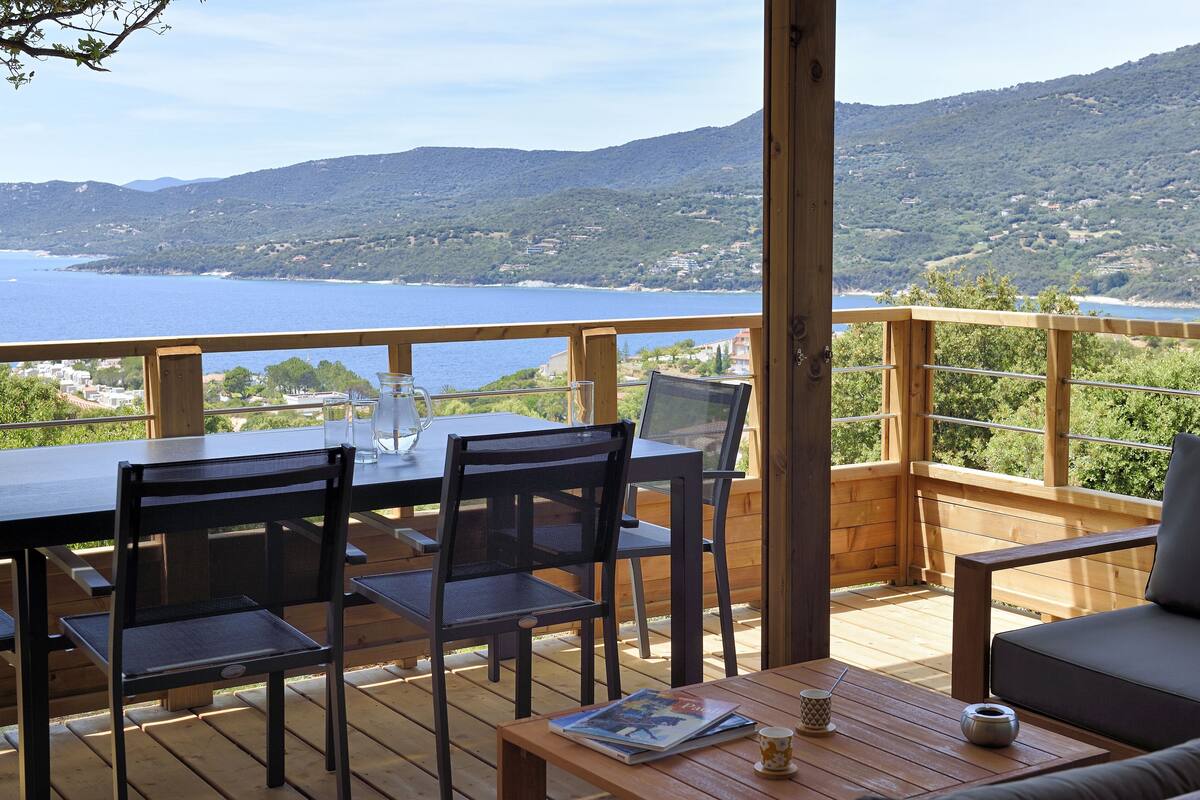 A wooden deck with a table and chairs, overlooking a body of water and hills.