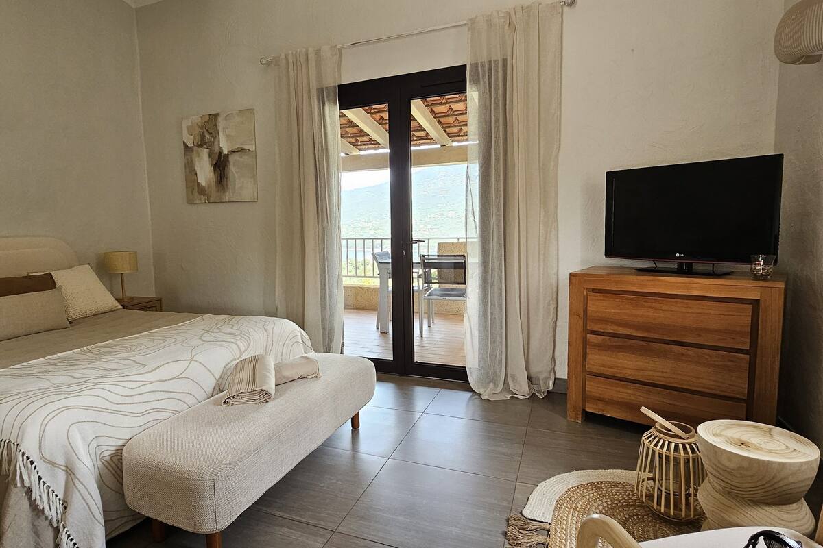 A bedroom with a bed, a TV on a wooden dresser, and a balcony with a view.