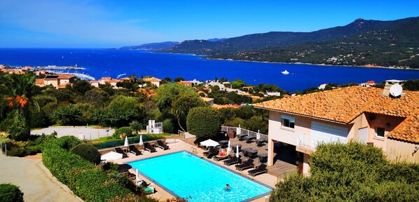 A pool area with lounge chairs and umbrellas, surrounded by greenery and overlooking a coastal view with buildings and a clear sky. - Residence & Hôtel Aria Marina (Viggianello)
