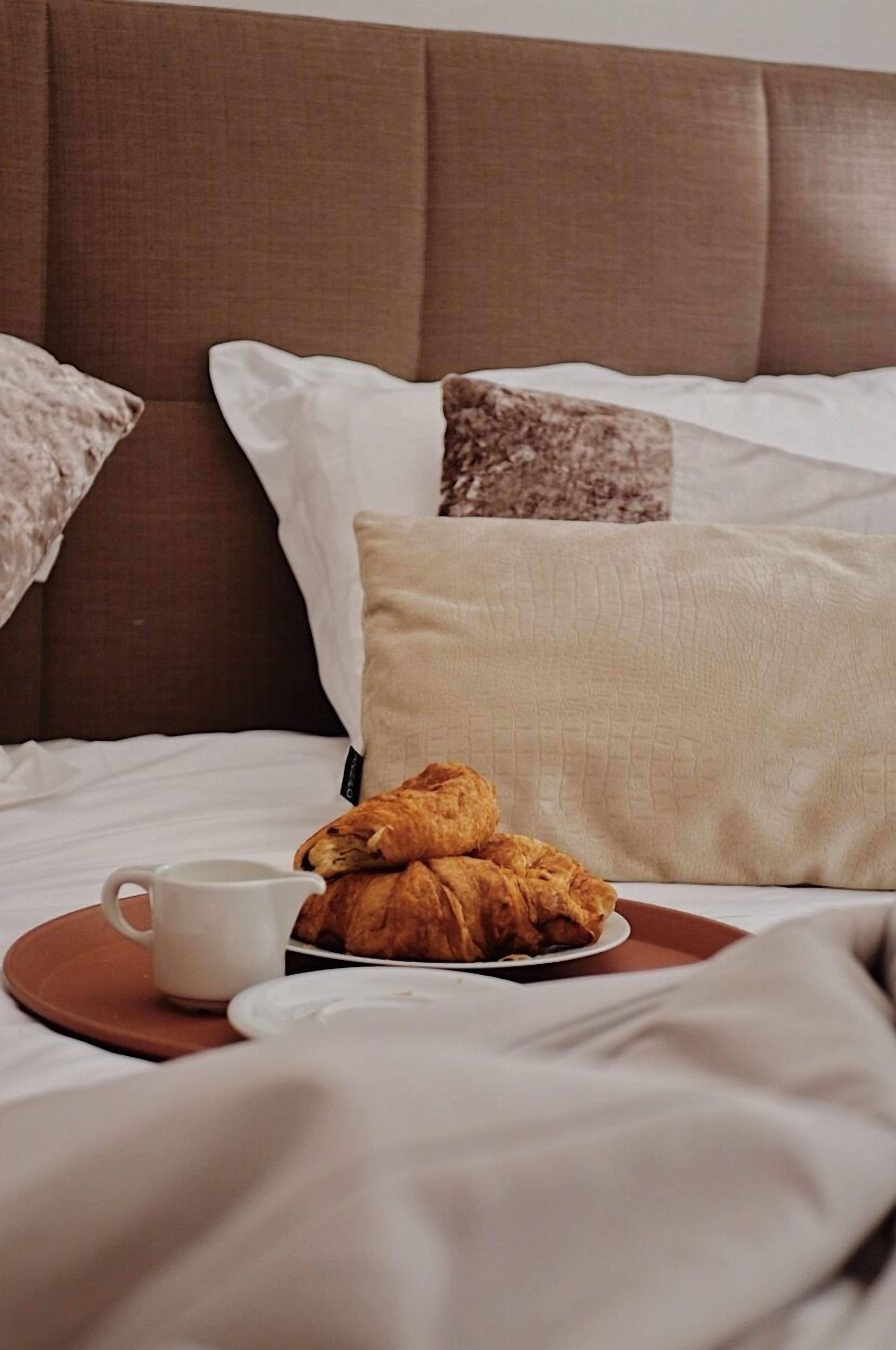 Een bed met een bruin hoofdeinde, witte kussens en een bord met croissants en een kop koffie.