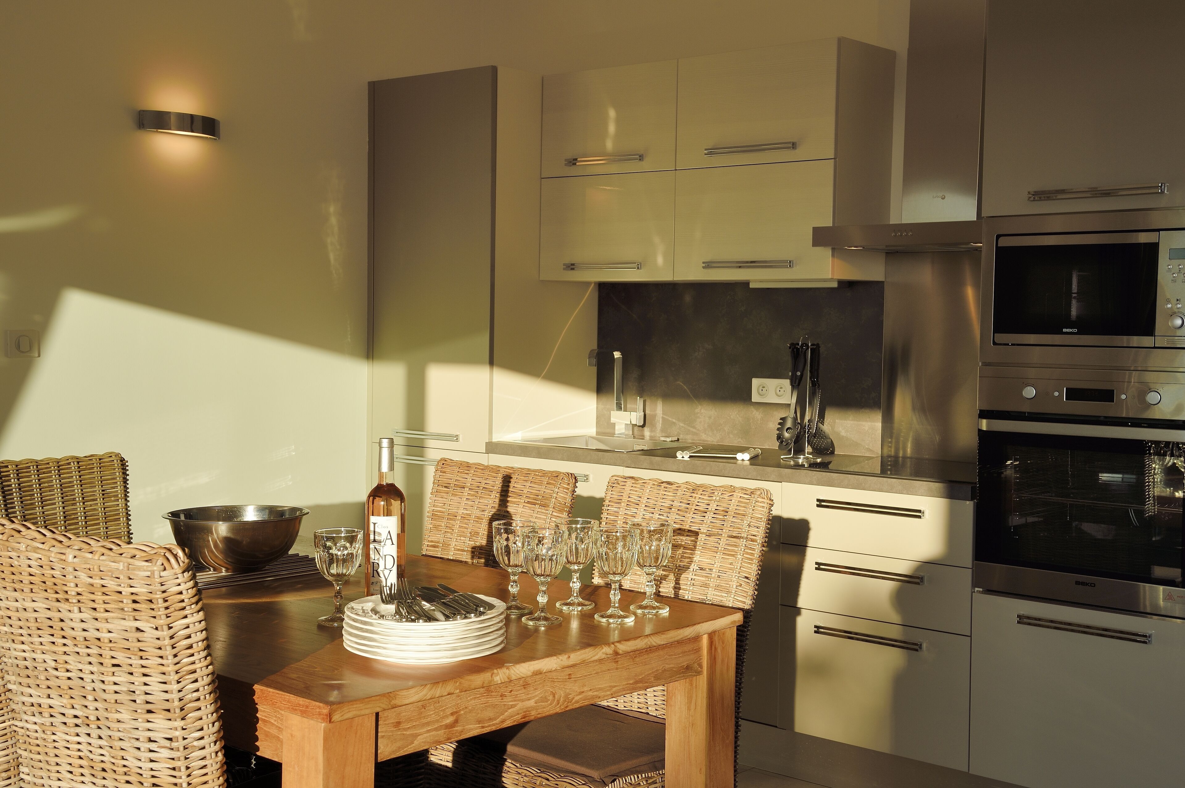 Una cocina moderna con electrodomésticos de acero inoxidable, una mesa de comedor con platos y vasos, y sillas de mimbre.