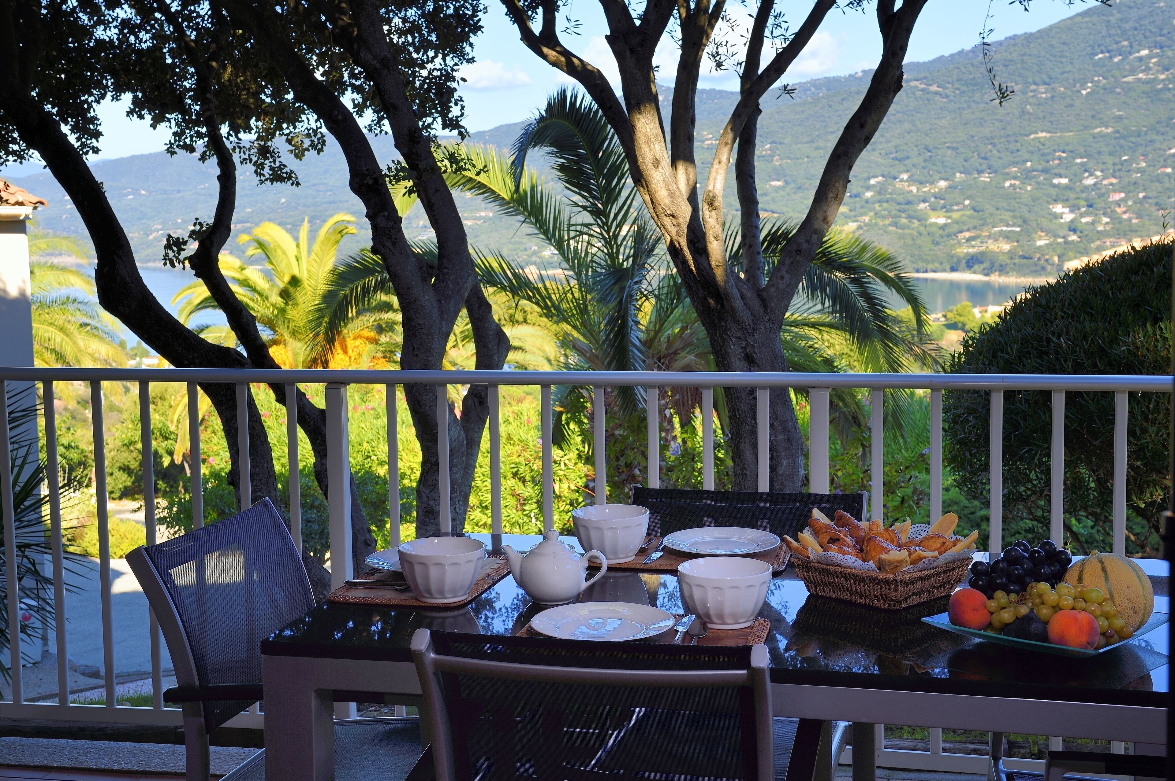 A table set for breakfast with white dishes, a teapot, and a fruit basket on a balcony overlooking a mountainous landscape.