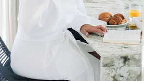 Une personne est assise à une table devant un livre, une tasse et une assiette contenant des croissants et du jus d’orange.