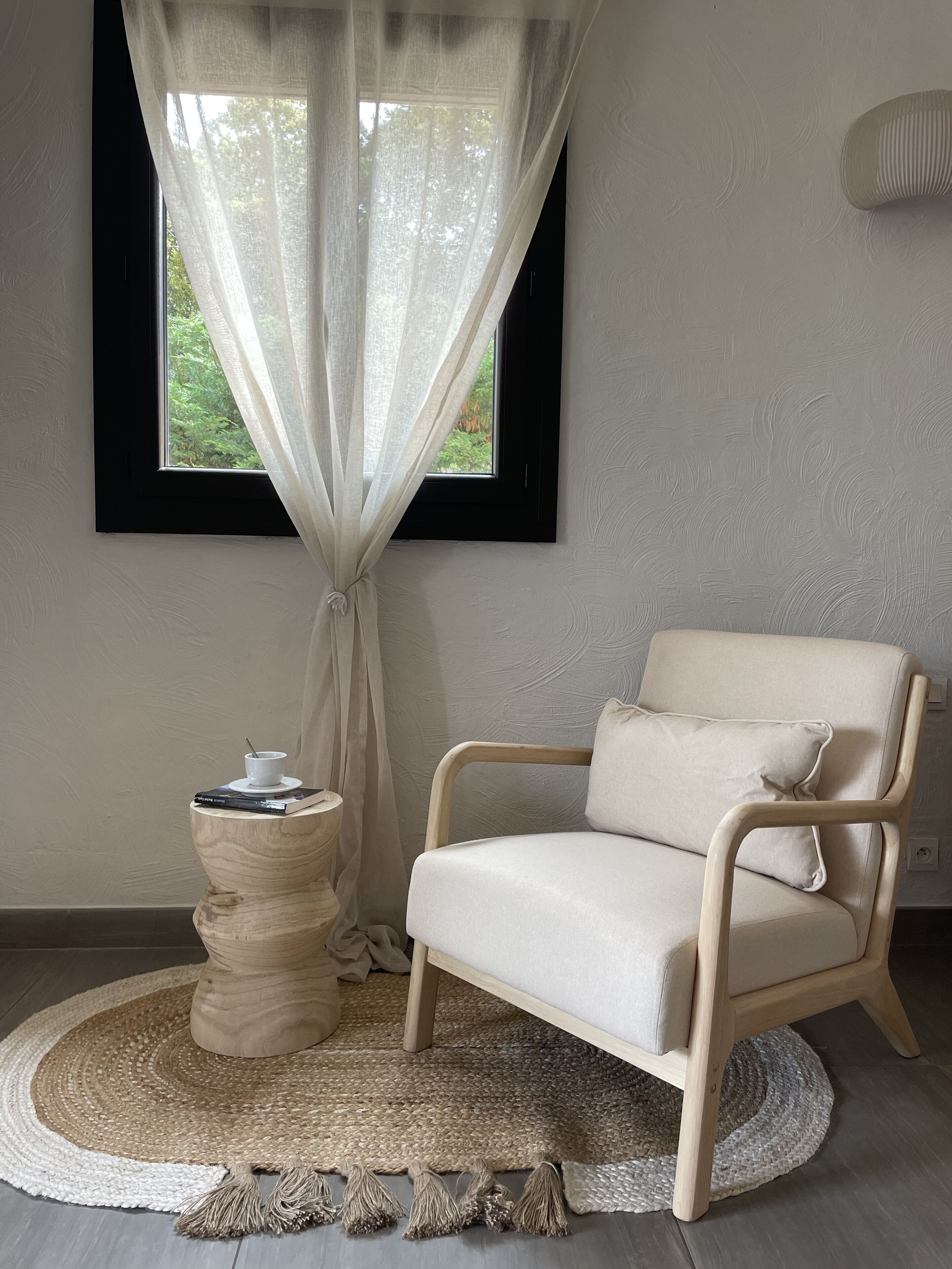 Ein Zimmer mit Fenster, einem Sessel, einem kleinen Tisch mit Tasse und Büchern sowie einem Teppich.