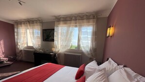 Une chambre d’hôtel avec un lit, un bureau, une chaise, un radiateur et une fenêtre avec des rideaux légers.