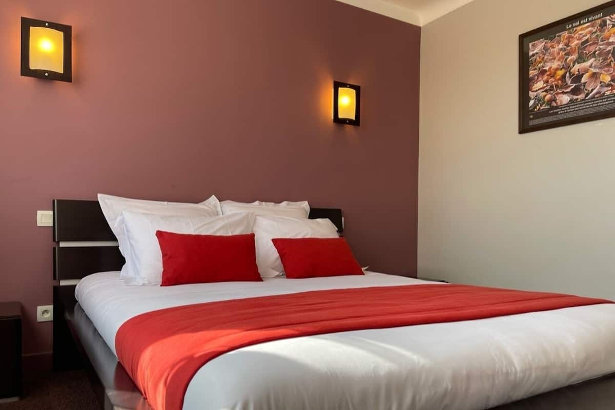 Una cama bien hecha con ropa de cama blanca y roja, dos luces fijadas a la pared y un cuadro enmarcado.